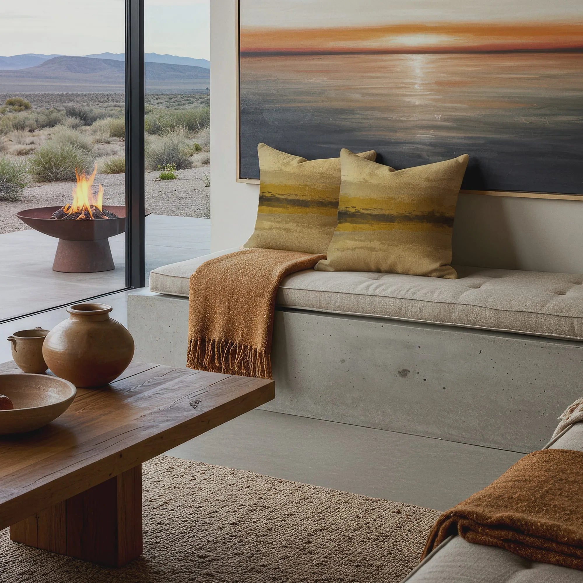 Modern desert living room with two abstract earth-toned throw pillows, tan throw blanket, rustic wood table, and outdoor fire pit view.