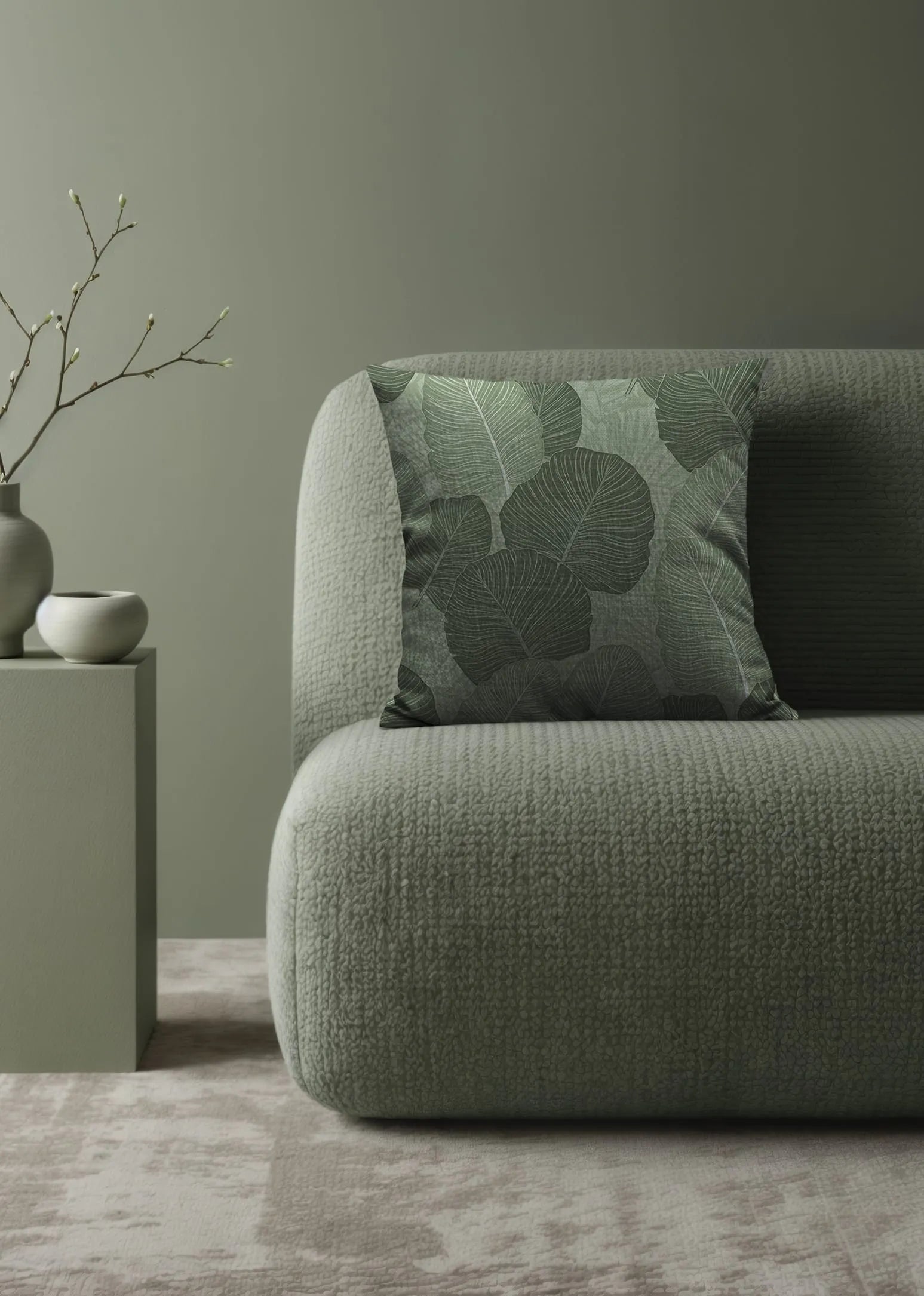 Minimalist green textured sofa with botanical leaf-patterned pillows beside white ceramic vases and a branch on a pedestal.