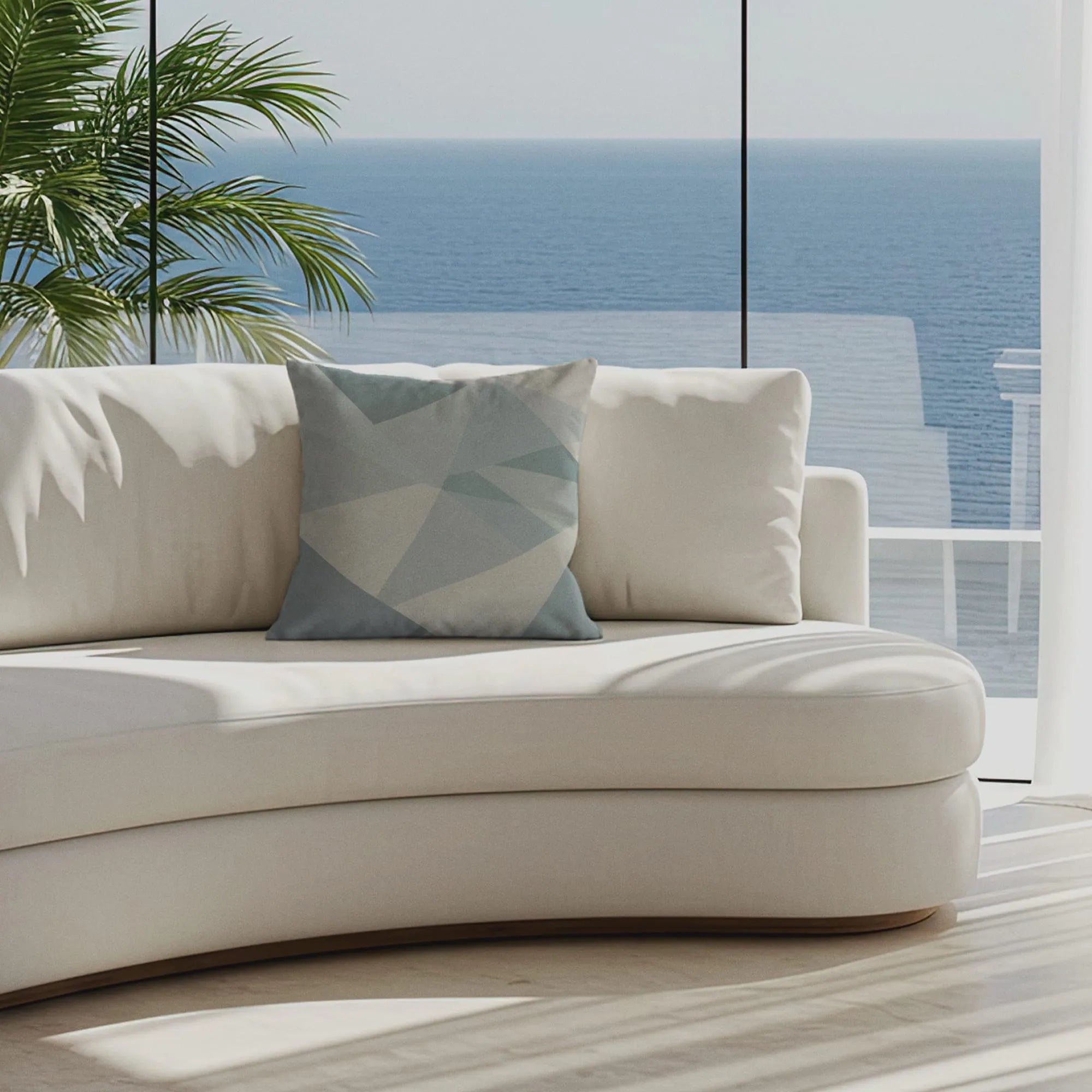 Modern coastal living room with curved white sofa, geometric blue Buffalo Bunny pillow, ocean view, and palm shadows through floor-to-ceiling windows.