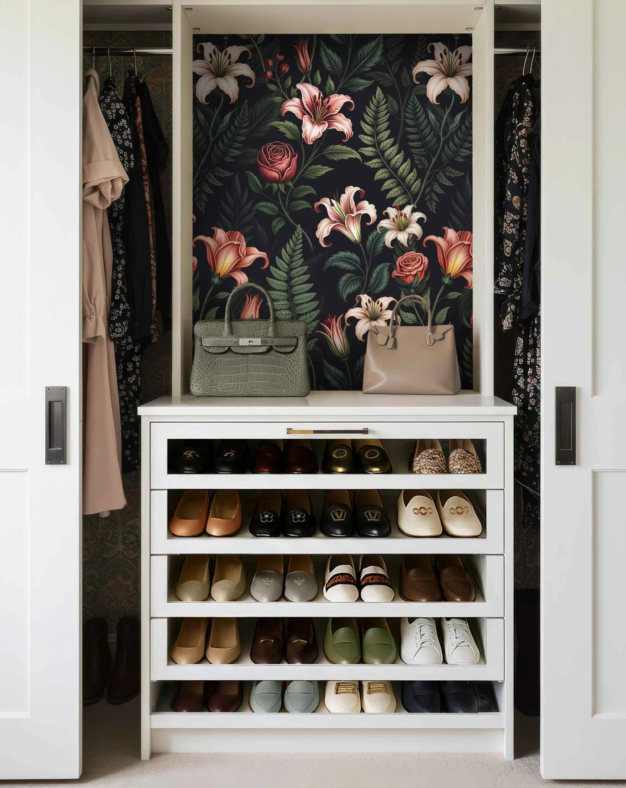 Closet filled with two handbags, cloths, a shoe rack, and floral pattern wallpaper with pink lilies, roses, green leaves on a dark background