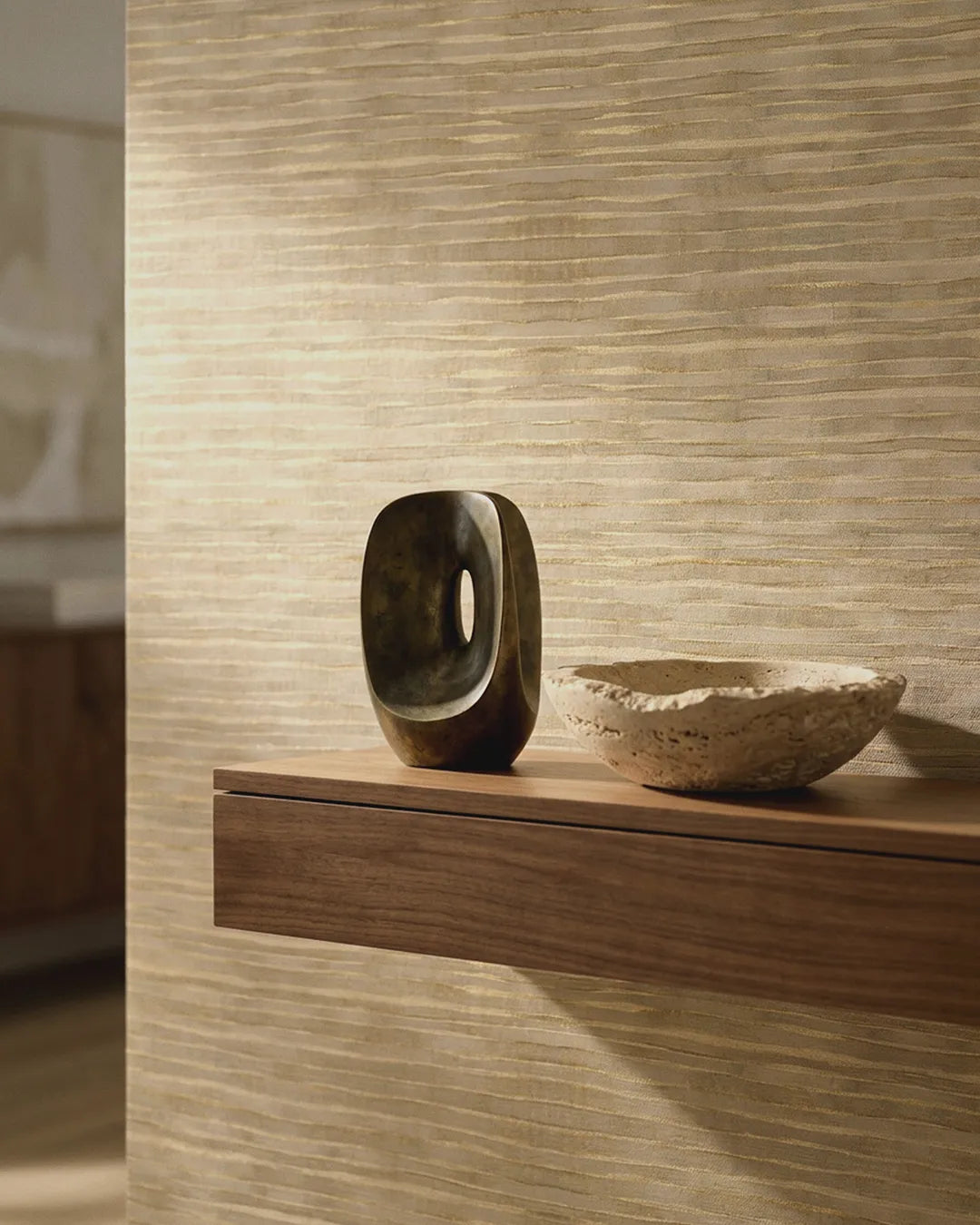 A modern floating wooden shelf mounted against a textured beige wall, holding a dark polished stone sculpture and a small rough stone bowl. The wall features the Ripple Weave Wallpaper.