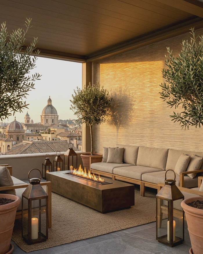 Outdoor patio seating area with a beige sectional sofa, rectangular fire pit table, woven rug, potted olive trees, and lanterns, overlooking a city skyline at sunset. The wall features the Ripple Weave Wallpaper.
