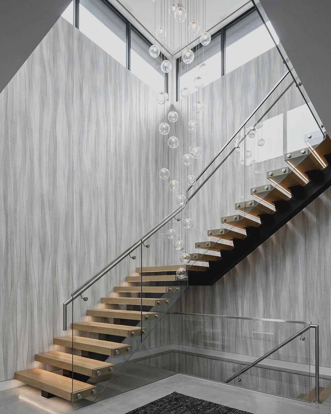 Modern floating staircase with light wood steps and clear glass railings, set in a contemporary interior with Soft Current Wallpaper walls and a hanging glass globe chandelier.