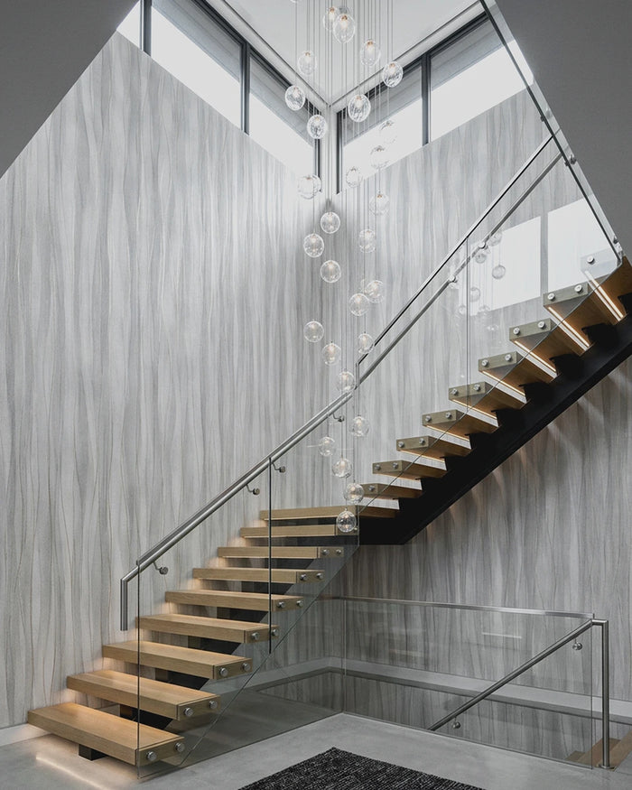 Modern floating staircase with light wood steps and clear glass railings, set in a contemporary interior with Soft Current Wallpaper walls and a hanging glass globe chandelier.
