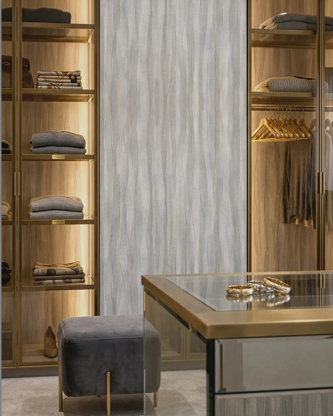 A luxury walk-in closet interior with wood shelving, gold-toned hardware, folded gray garments, hanging clothes, and a gray velvet ottoman in front of a glass-top vanity. The middle wall features the grey Soft Current Wallpaper.