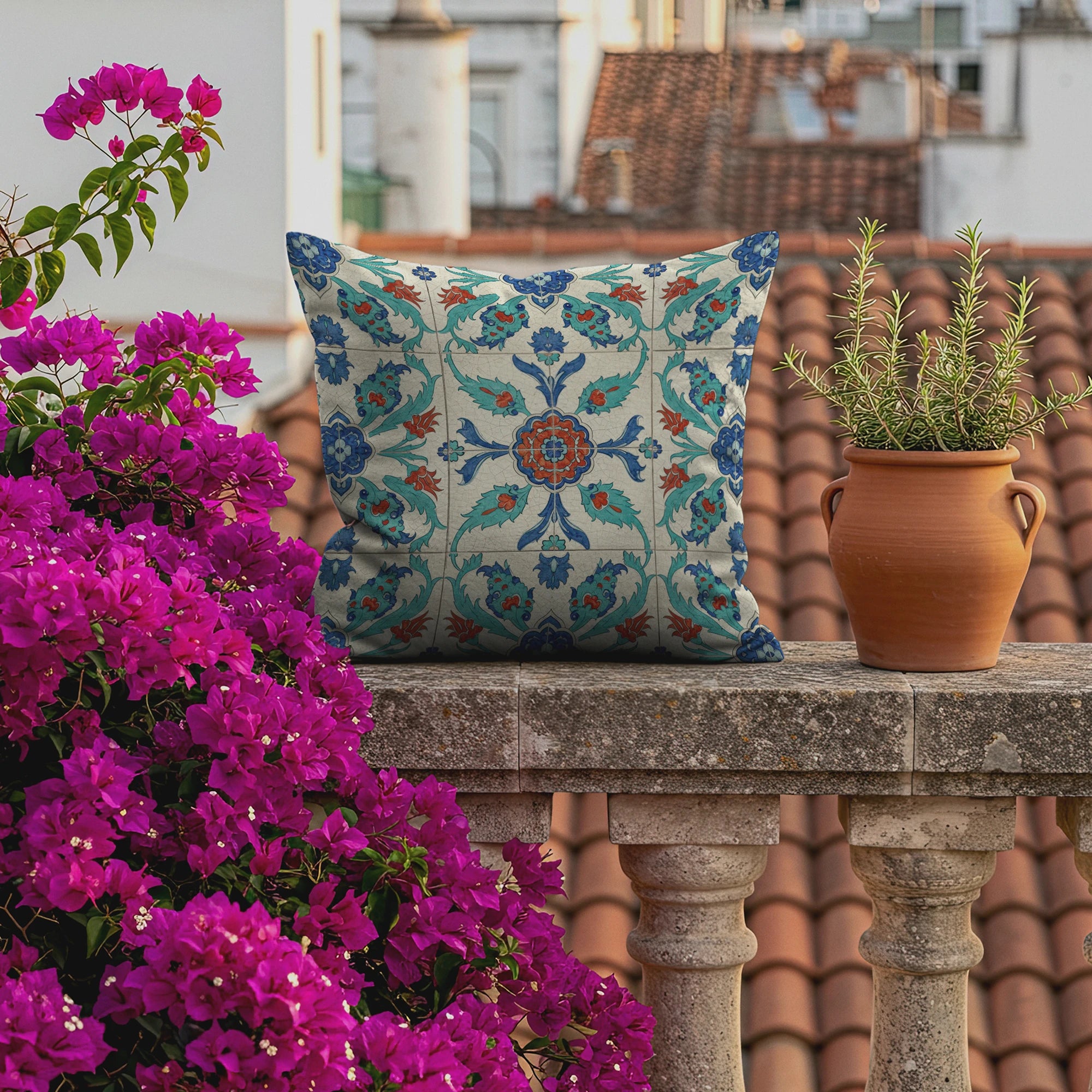 European rooftop terrace scene featuring a boho tile-print pillow in teal, blue, and rust hues, lush bougainvillea flowers, terracotta pot with rosemary, and charming city rooftops in the background.