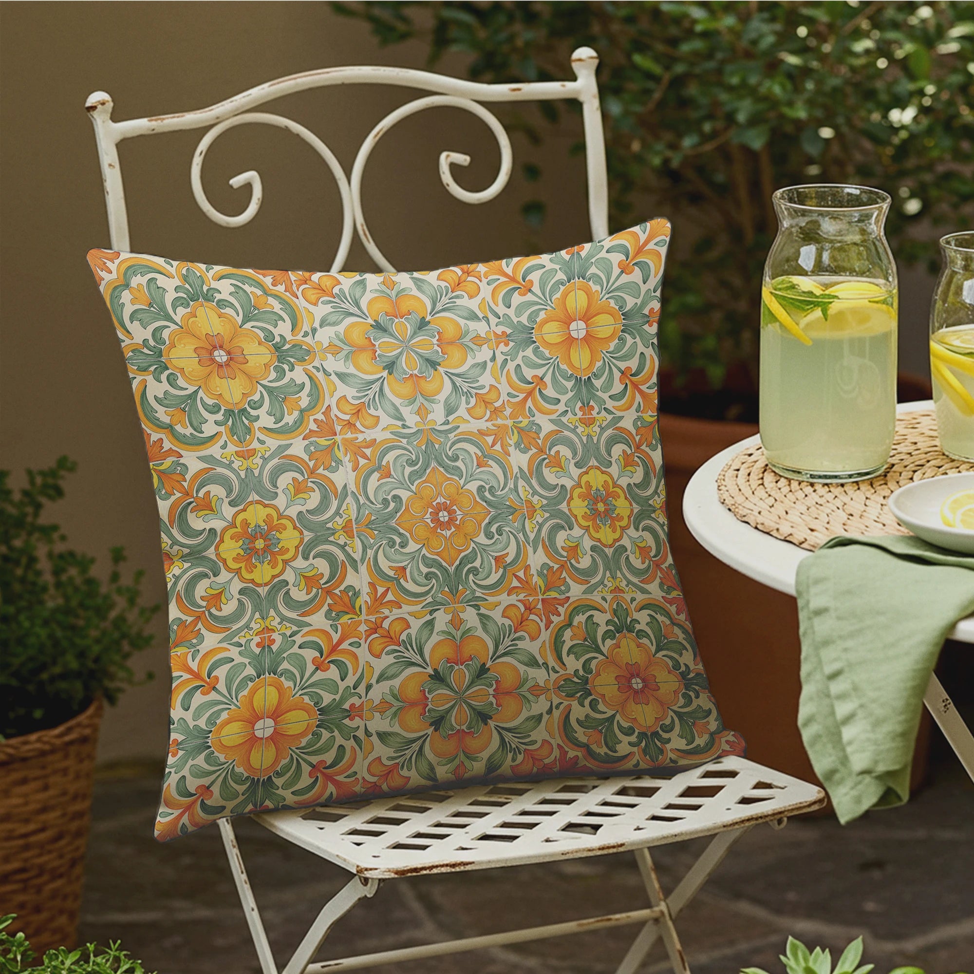 Outdoor bistro setting with white metal chair, vibrant floral tile-print pillow, pitcher of lemonade, and round table with a green napkin.