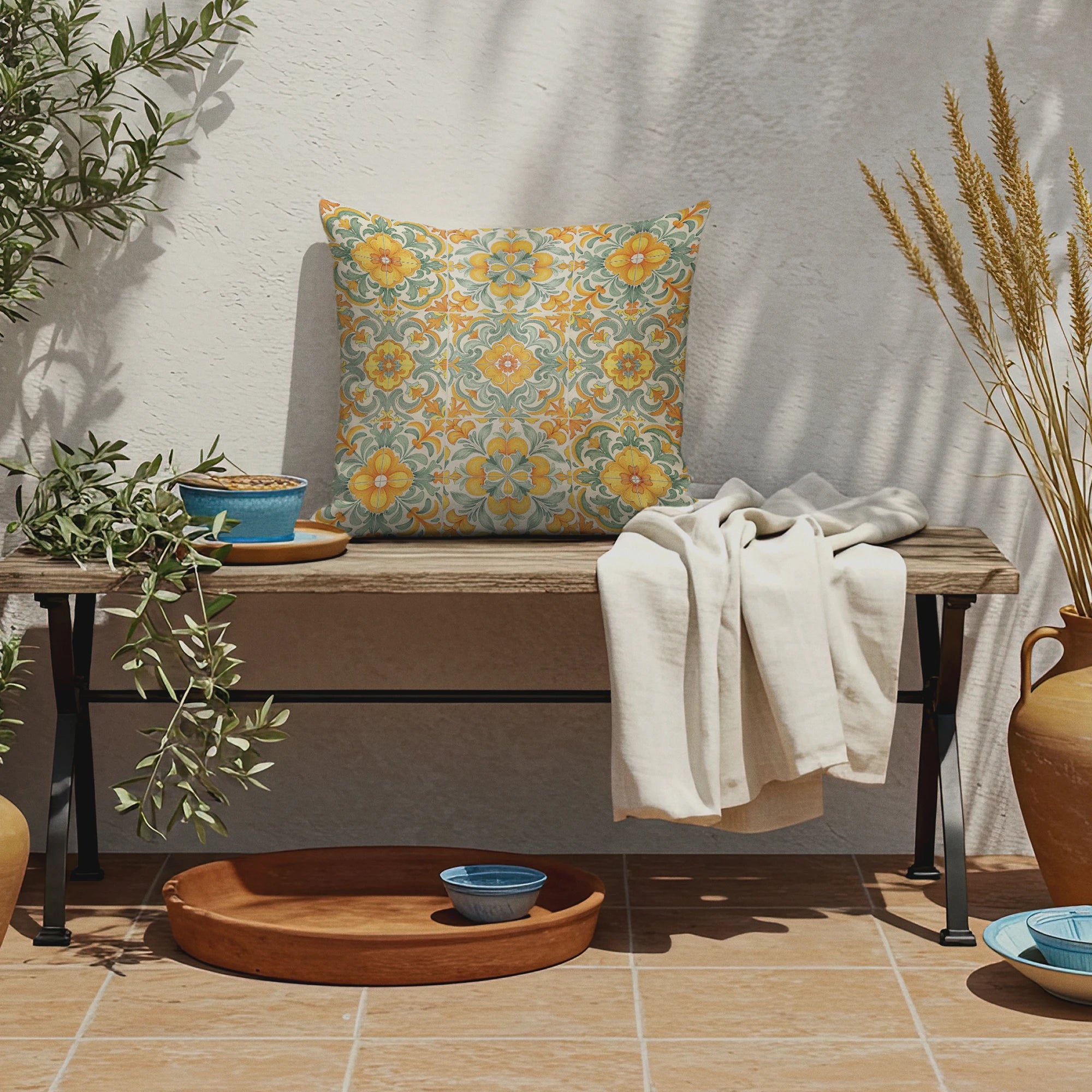 Mediterranean patio bench with vibrant floral tile-print pillow, olive branches, ceramic bowls, terracotta tray, and neutral throw blanket.